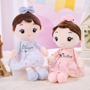 May include: Two plush dolls with brown hair and bows, one blue and one pink. The blue doll has the name "Morgan" on its dress, and the pink doll has the name "Destiny". Both dolls have light skin and are wearing dresses.