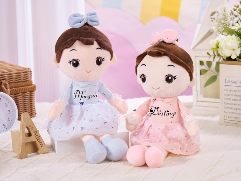 May include: Two plush dolls with brown hair and bows, one blue and one pink. The blue doll has the name "Morgan" on its dress, and the pink doll has the name "Destiny". Both dolls have light skin and are wearing dresses.