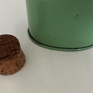 May include: A brown cork bottle stopper next to a light green metal container. The cork has a textured surface with visible grain. The container has a black rim and a slightly distressed finish.