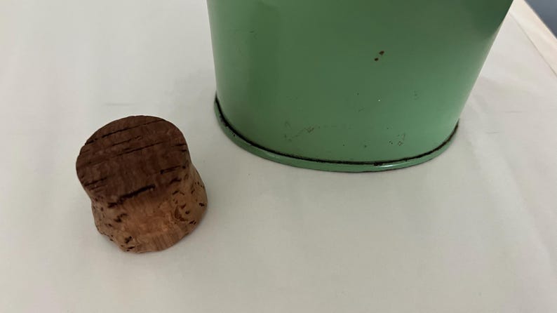 May include: A brown cork bottle stopper next to a light green metal container. The cork has a textured surface with visible grain. The container has a black rim and a slightly distressed finish.