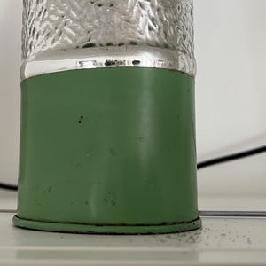May include: A vintage-style lamp with a green base and a textured, clear glass top. The base is cylindrical and has a silver band at the top. The lamp is on a white surface.