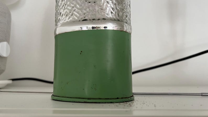 May include: A vintage-style lamp with a green base and a textured, clear glass top. The base is cylindrical and has a silver band at the top. The lamp is on a white surface.