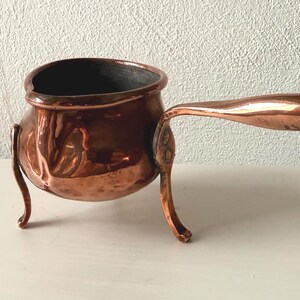 May include: A small, antique copper pot with a long handle and three decorative legs. The pot has a rounded body with embossed floral designs and a dark interior. The handle is curved and extends horizontally from the pot's side.