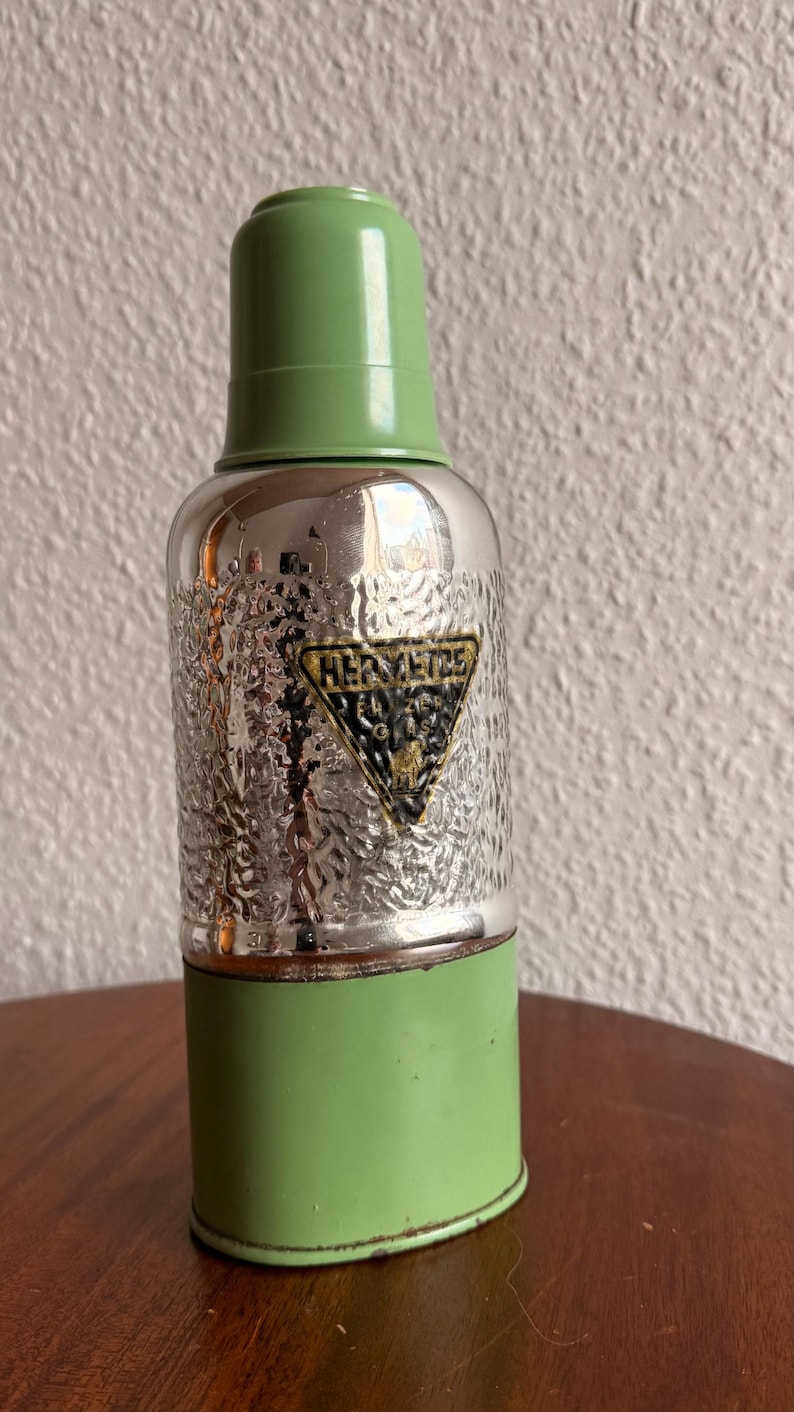 May include: Vintage thermos with a textured silver body, green base, and lid. The thermos features a black and gold emblem that reads "HERMETICS". A classic, retro item.