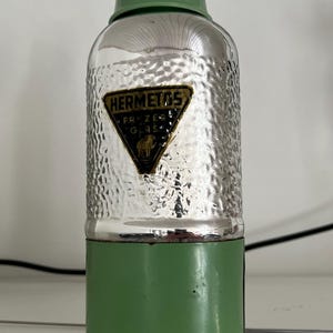 May include: Vintage green and silver metal bottle with a textured surface. The bottle has a green top and base, with a silver hammered-look center. A black and gold label reads "HERMETOS".