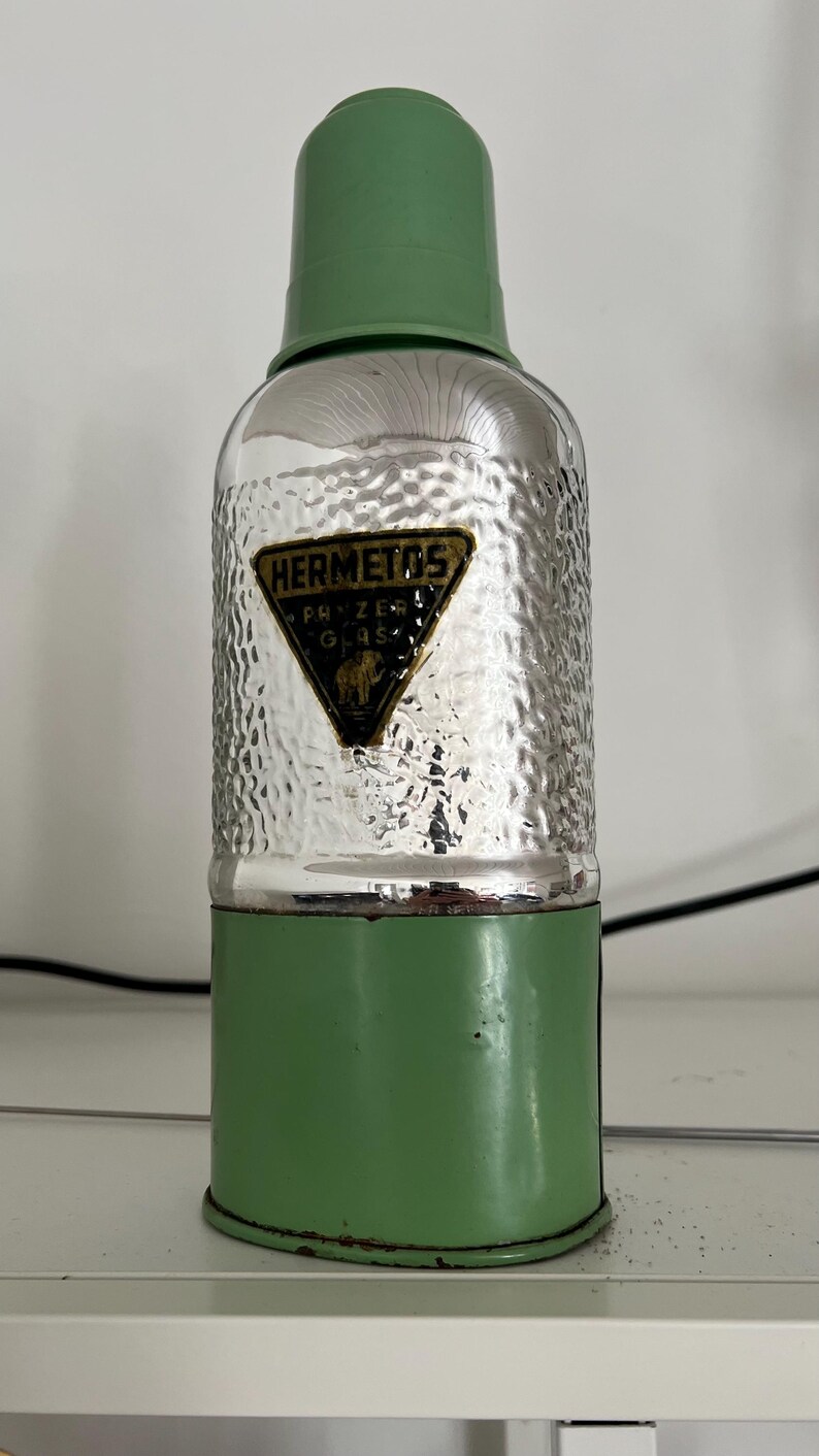 May include: Vintage green and silver metal bottle with a textured surface. The bottle has a green top and base, with a silver hammered-look center. A black and gold label reads "HERMETOS".
