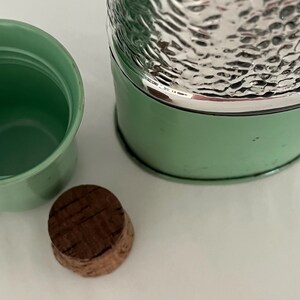 May include: A vintage green thermos with a silver textured top, a matching green cup, and a cork stopper. The thermos has a silver rim and a light green body. The cup and thermos are empty.