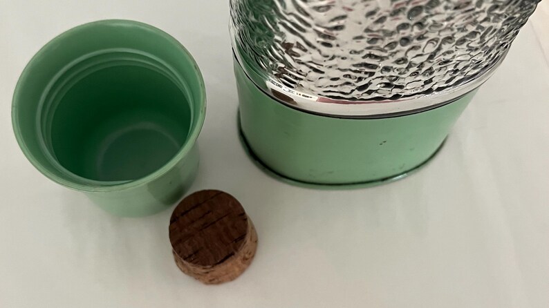 May include: A vintage green thermos with a silver textured top, a matching green cup, and a cork stopper. The thermos has a silver rim and a light green body. The cup and thermos are empty.