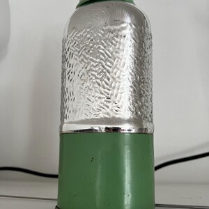 May include: A vintage-style green and silver metal bottle. The top and bottom sections are green, while the middle section has a textured silver finish. The bottle is cylindrical with a rounded top.