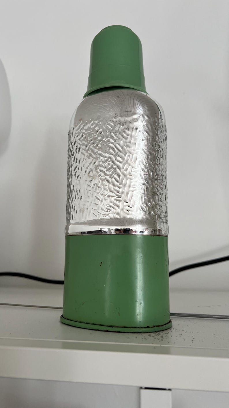 May include: A vintage-style green and silver metal bottle. The top and bottom sections are green, while the middle section has a textured silver finish. The bottle is cylindrical with a rounded top.