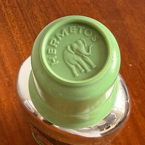 May include: A green bottle stopper with the word "HERMETOS" and an elephant design embossed on the top. The stopper is set on a clear glass bottle with a silver-colored metal top. The background is a wooden surface.