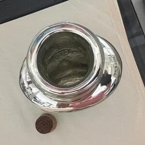 May include: A silver-colored, open-top container with a wide, rounded base and a small, round, wooden stopper. The container reflects the surrounding environment. The item is likely a decorative or functional object.