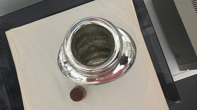 May include: A silver-colored, open-top container with a wide, rounded base and a small, round, wooden stopper. The container reflects the surrounding environment. The item is likely a decorative or functional object.