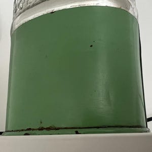 May include: A vintage green metal container with a clear glass top. The container has a cylindrical shape and shows signs of wear, including rust at the base. The background is a white wall and a gray speaker.