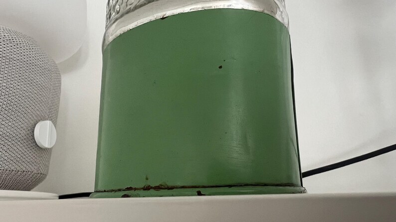 May include: A vintage green metal container with a clear glass top. The container has a cylindrical shape and shows signs of wear, including rust at the base. The background is a white wall and a gray speaker.