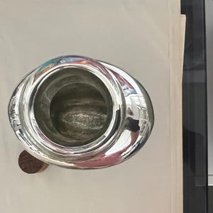 May include: A shiny, silver-colored metal pitcher with a wide, oval-shaped body and a wooden base. The pitcher's interior is visible, and the surface reflects the surrounding environment. The design is simple and elegant.