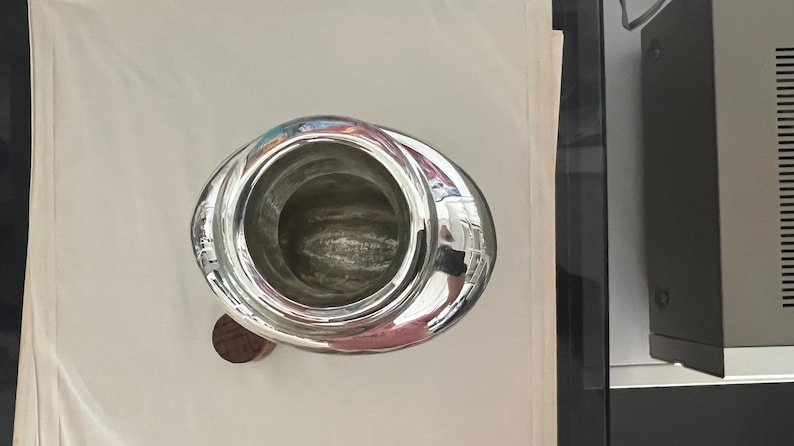 May include: A shiny, silver-colored metal pitcher with a wide, oval-shaped body and a wooden base. The pitcher's interior is visible, and the surface reflects the surrounding environment. The design is simple and elegant.