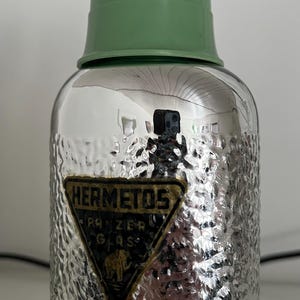 May include: Vintage glass thermos with a textured silver body and a green plastic lid. The thermos features a label with the text "HERMETOS" and "PANZER GLAS" with an elephant illustration. A classic item for hot or cold beverages.
