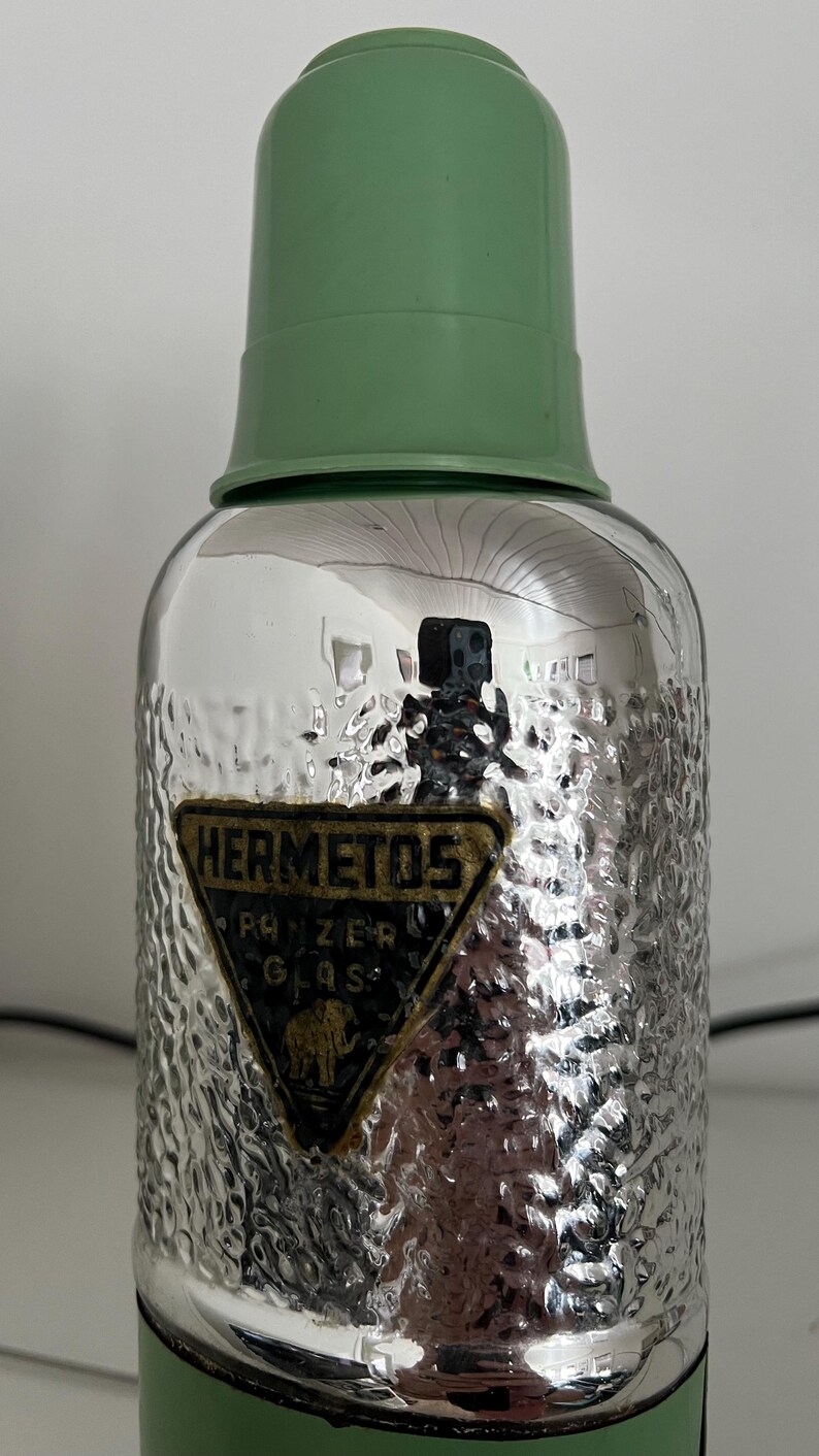 May include: Vintage glass thermos with a textured silver body and a green plastic lid. The thermos features a label with the text "HERMETOS" and "PANZER GLAS" with an elephant illustration. A classic item for hot or cold beverages.