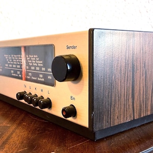 Könnte beinhalten: Vintage Radioempfänger mit silberner Front und Holzmaserung. Es hat schwarze Knöpfe, ein Wahlrad und das Wort "Sender". Das Radio steht auf einer Holzoberfläche.