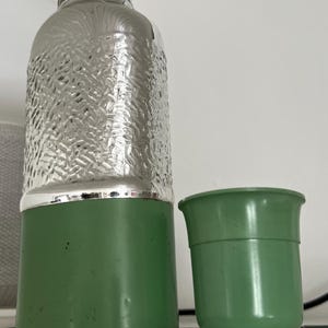 May include: A vintage-style green and silver water bottle with a matching green cup. The bottle has a textured silver upper half and a green base. The cup is a solid green color.