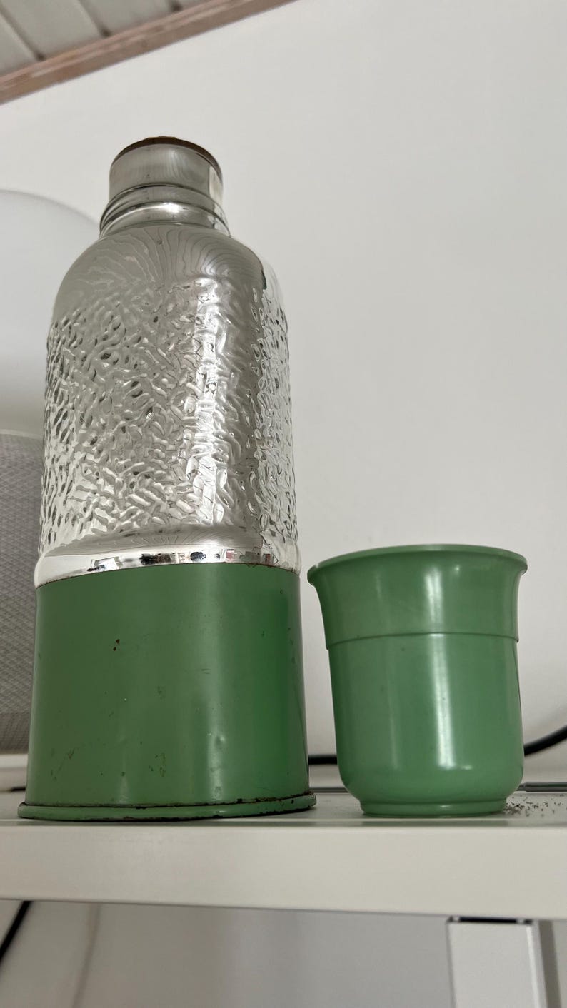 May include: A vintage-style green and silver water bottle with a matching green cup. The bottle has a textured silver upper half and a green base. The cup is a solid green color.