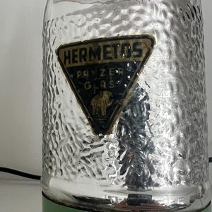 May include: Vintage green and silver glass thermos. The thermos has a textured silver body and a green top and base. A triangular label reads "HERMETOS" with "PANZER GLAS" and an elephant illustration.