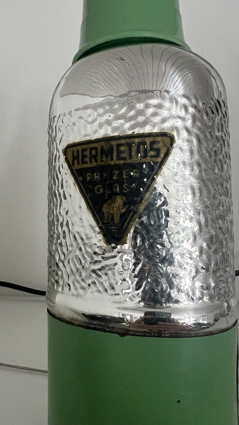 May include: Vintage green and silver glass thermos. The thermos has a textured silver body and a green top and base. A triangular label reads "HERMETOS" with "PANZER GLAS" and an elephant illustration.