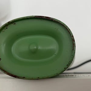 May include: A green, oval-shaped metal object with a slightly distressed finish. The object has a central indentation and a dark, possibly rusted, rim. A black cord extends from the side. The object is likely a component of a lamp or similar device.