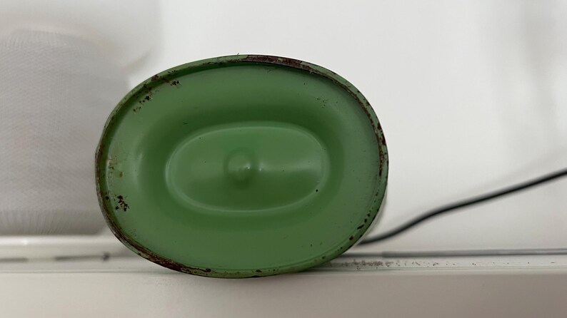 May include: A green, oval-shaped metal object with a slightly distressed finish. The object has a central indentation and a dark, possibly rusted, rim. A black cord extends from the side. The object is likely a component of a lamp or similar device.