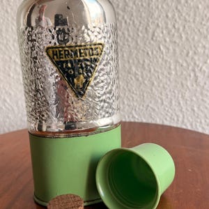 May include: Vintage silver-toned flask with a textured surface and a green base. The flask has a green cup and a cork stopper. The flask has a gold and black label that reads "HERMETOS".