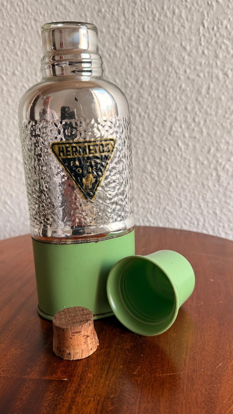 May include: Vintage silver-toned flask with a textured surface and a green base. The flask has a green cup and a cork stopper. The flask has a gold and black label that reads "HERMETOS".