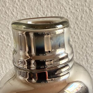 May include: Close-up of a clear glass bottle with a silver-colored neck and rim. The bottle's surface reflects light, creating a shimmering effect. The design features a rounded body and a slightly flared neck, suggesting a decorative or functional item.