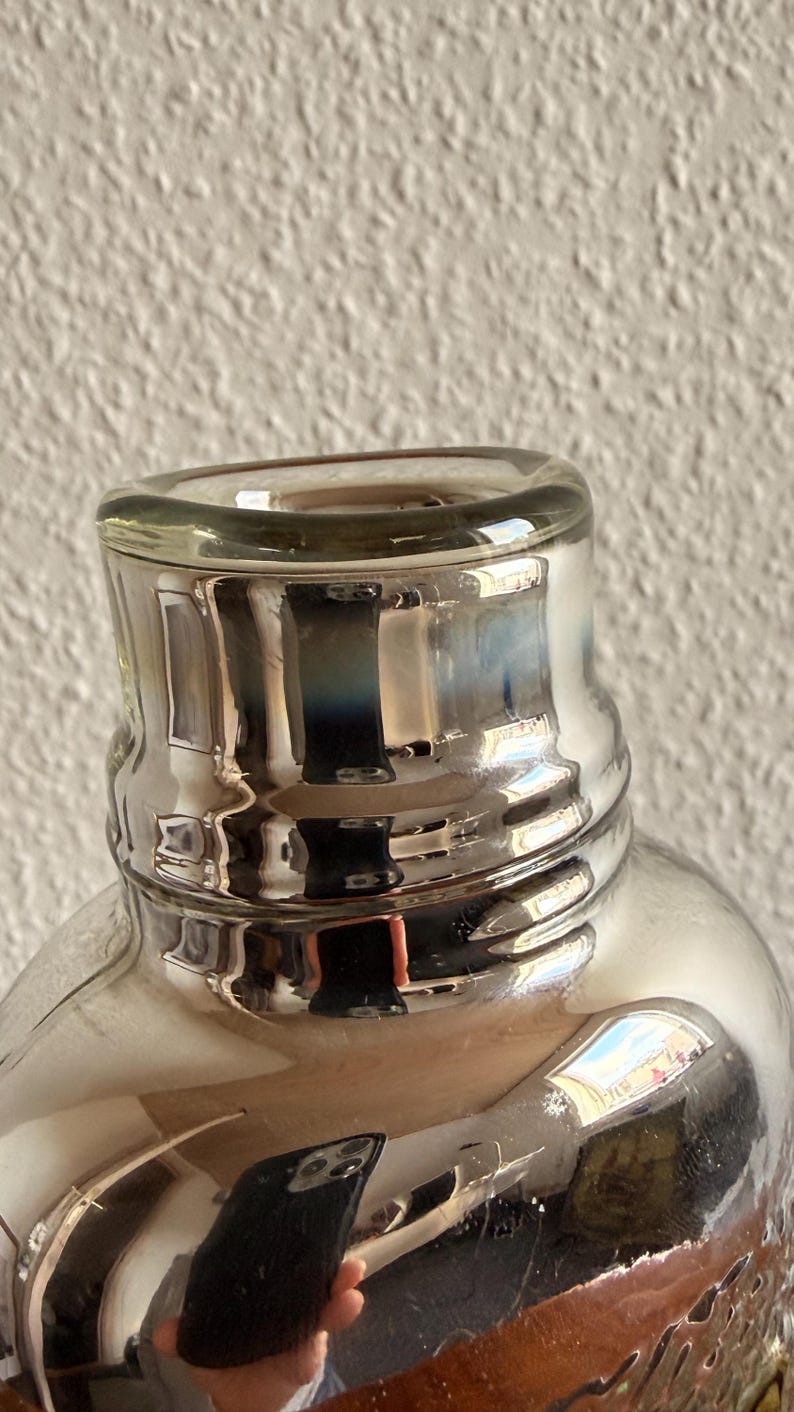 May include: Close-up of a clear glass bottle with a silver-colored neck and rim. The bottle's surface reflects light, creating a shimmering effect. The design features a rounded body and a slightly flared neck, suggesting a decorative or functional item.
