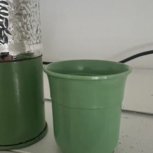 May include: A light green, vintage-style planter with a rounded shape. The planter is made of a solid material and has a smooth, matte finish. A silver, textured object is visible in the background. The planter is likely for indoor or outdoor use.