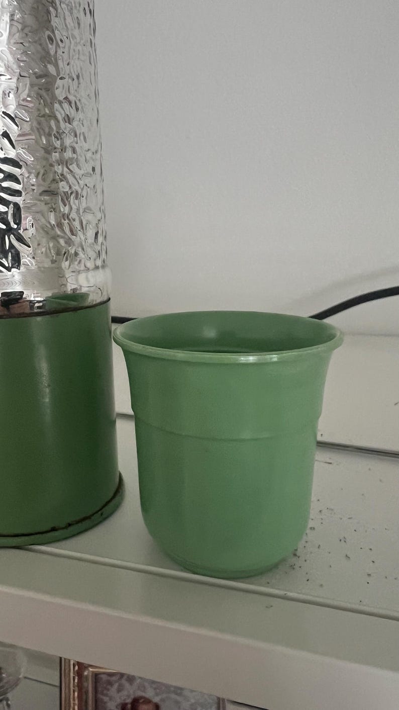 May include: A light green, vintage-style planter with a rounded shape. The planter is made of a solid material and has a smooth, matte finish. A silver, textured object is visible in the background. The planter is likely for indoor or outdoor use.