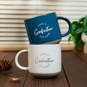 Puede incluir: Dos tazas de cerámica, una turquesa y otra blanca, apiladas sobre una superficie de madera. La taza turquesa, con la inscripción "Luke's Godfather EST. 2025", está encima de la taza blanca, que dice "Luke's Godmother EST. 2025". Ambas tazas tienen una base gris moteada.
