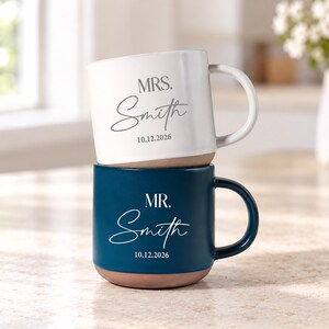 Puede incluir: Una taza de cerámica blanca apilada sobre una taza de cerámica azul oscuro. La taza blanca presenta el texto MRS. Smith y 10.12.2026 en gris. La taza azul presenta el texto MR. Smith y 10.12.2026 en blanco. Ambas tazas tienen una base de arcilla.