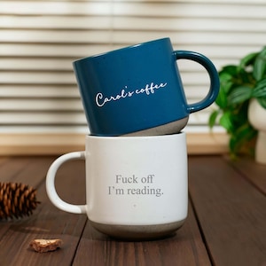 May include: A stack of two ceramic mugs with a speckled finish. The top mug is dark blue with the white script text 'Carol's coffee'. The bottom mug is white with the grey text 'Fuck off I'm reading.' Both mugs feature a smooth, unglazed base.