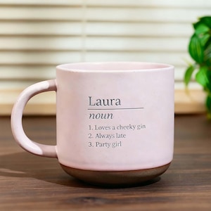May include: A pink ceramic mug with a brown base and a curved handle. The mug features the name "Laura" with the definition "noun" and three descriptive statements: "Loves a cheeky gin," "Always late," and "Party girl."