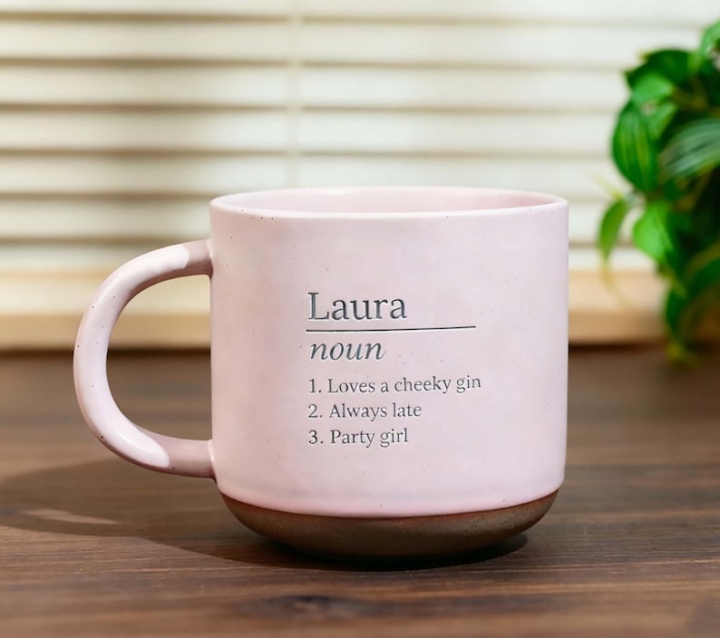 May include: A pink ceramic mug with a brown base and a curved handle. The mug features the name "Laura" with the definition "noun" and three descriptive statements: "Loves a cheeky gin," "Always late," and "Party girl."