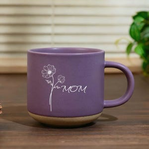 Pode incluir: Uma caneca de cerâmica roxa com base castanha clara. A caneca apresenta um design floral branco e a palavra "MOM" em escrita cursiva. A caneca está sobre uma superfície de madeira.