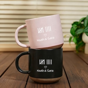 Könnte beinhalten: Zwei Keramikbecher, einer rosa und einer schwarz, übereinander gestapelt. Jeder Becher hat ein weißes Design mit vertikalen Linien, einem Herz und den Namen "Noah & Sara". Die Becher haben einen braunen Boden und einen gebogenen Henkel.