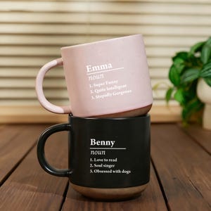May include: Two ceramic mugs stacked on top of each other. The top mug is pink with the name "Emma" and the definition of a noun, with three descriptive qualities. The bottom mug is black with the name "Benny" and the definition of a noun, with three descriptive qualities.