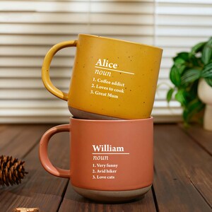 May include: Two ceramic mugs stacked on top of each other. The top mug is mustard yellow with the name "Alice" and the definition of "noun" printed in white, along with three descriptive phrases. The bottom mug is coral with the name "William" and the definition of "noun" printed in white, along with three descriptive phrases.