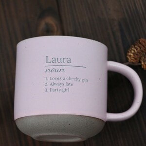 May include: A light pink ceramic mug with a gray base and handle. The mug has the name "Laura" printed on it, along with the word "noun" and three descriptive phrases: "Loves a cheeky gin," "Always late," and "Party girl."