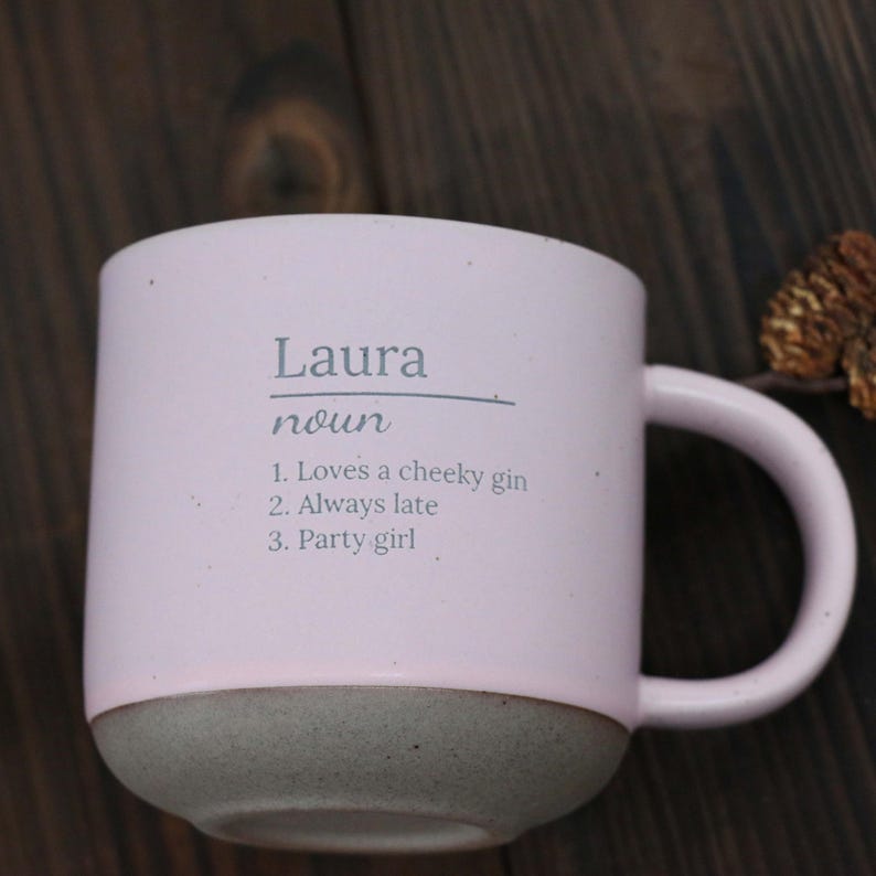 May include: A light pink ceramic mug with a gray base and handle. The mug has the name "Laura" printed on it, along with the word "noun" and three descriptive phrases: "Loves a cheeky gin," "Always late," and "Party girl."