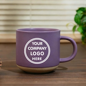 May include: A purple ceramic mug with a rounded handle and a tan base. The mug features the text "YOUR COMPANY LOGO HERE" in a white circular design. The mug is on a wooden surface.