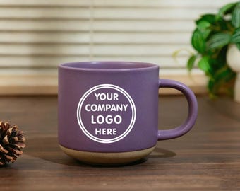 May include: A purple ceramic mug with a rounded handle and a tan base. The mug features the text "YOUR COMPANY LOGO HERE" in a white circular design. The mug is on a wooden surface.