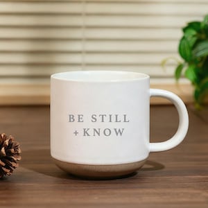 May include: White ceramic mug with the text "BE STILL + KNOW" in gray lettering. The mug has a brown base and a curved handle. A pinecone sits to the left of the mug.
